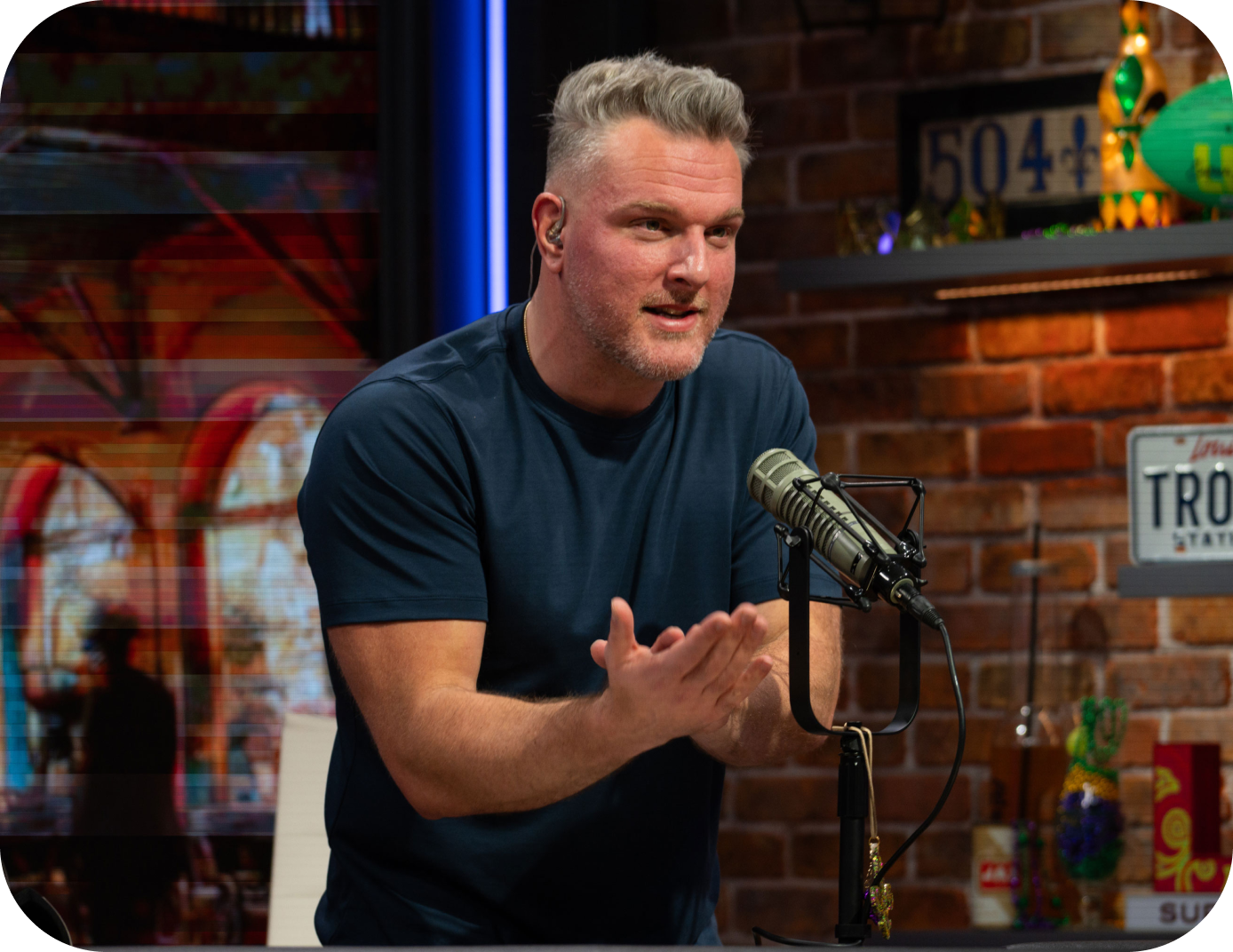 Pat McAfee speaking into a microphone in a studio with brick walls and colorful decorations.